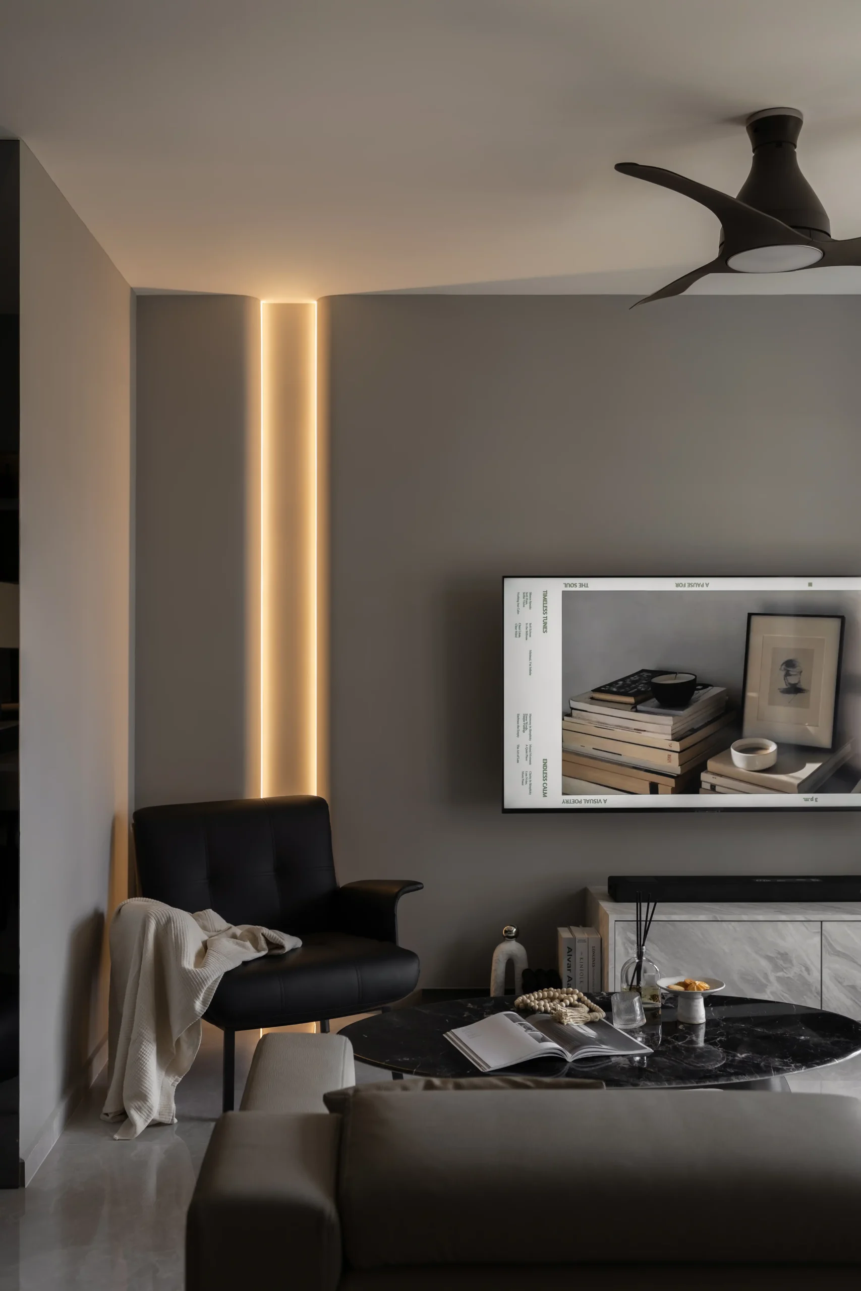 dark luxury interior living room featuring a recessed warm vertical light wall accent, a sleek black armchair, and a wall-mounted TV.