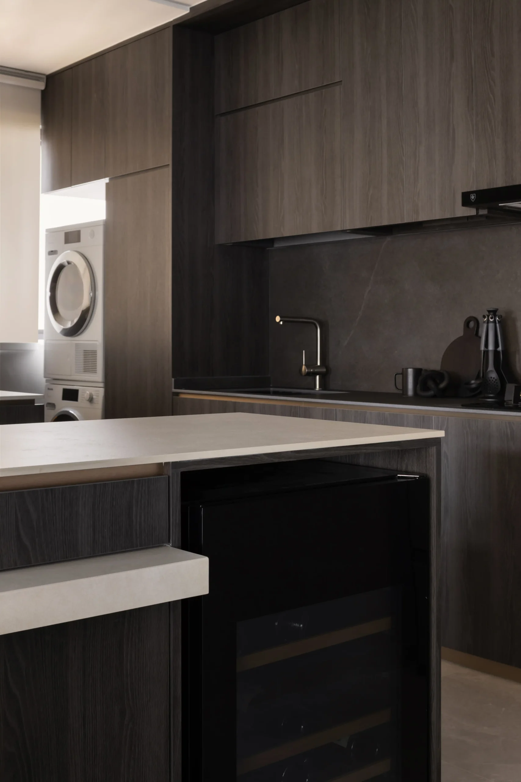 close-up of a contemporary moody kitchen island clad in dark wood grain laminate, featuring a built-in black glass wine cooler fridge.