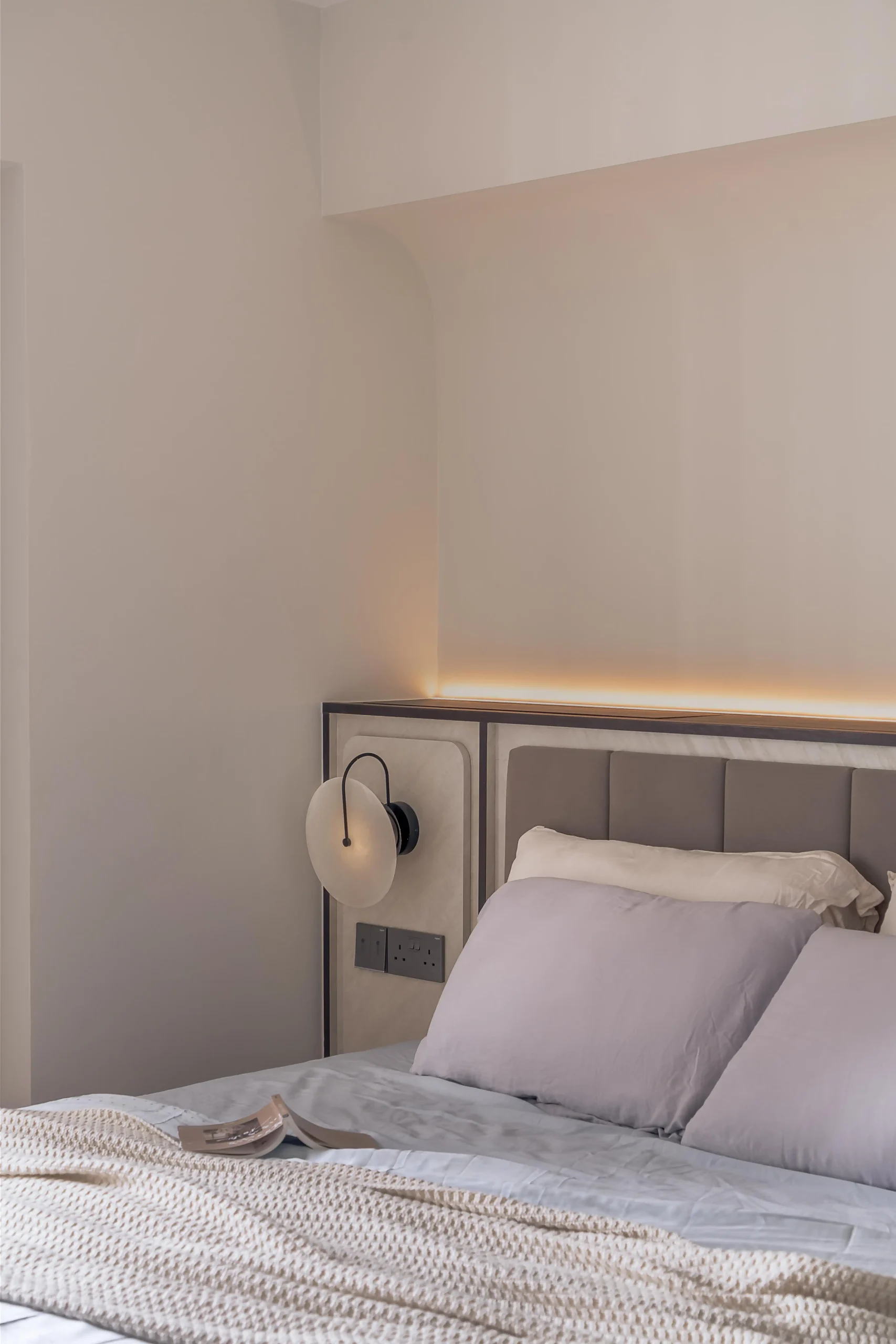 Master bedroom headboard niche with warm LED cove lighting, upholstered panel, and circular frosted glass wall sconce in modern interior design