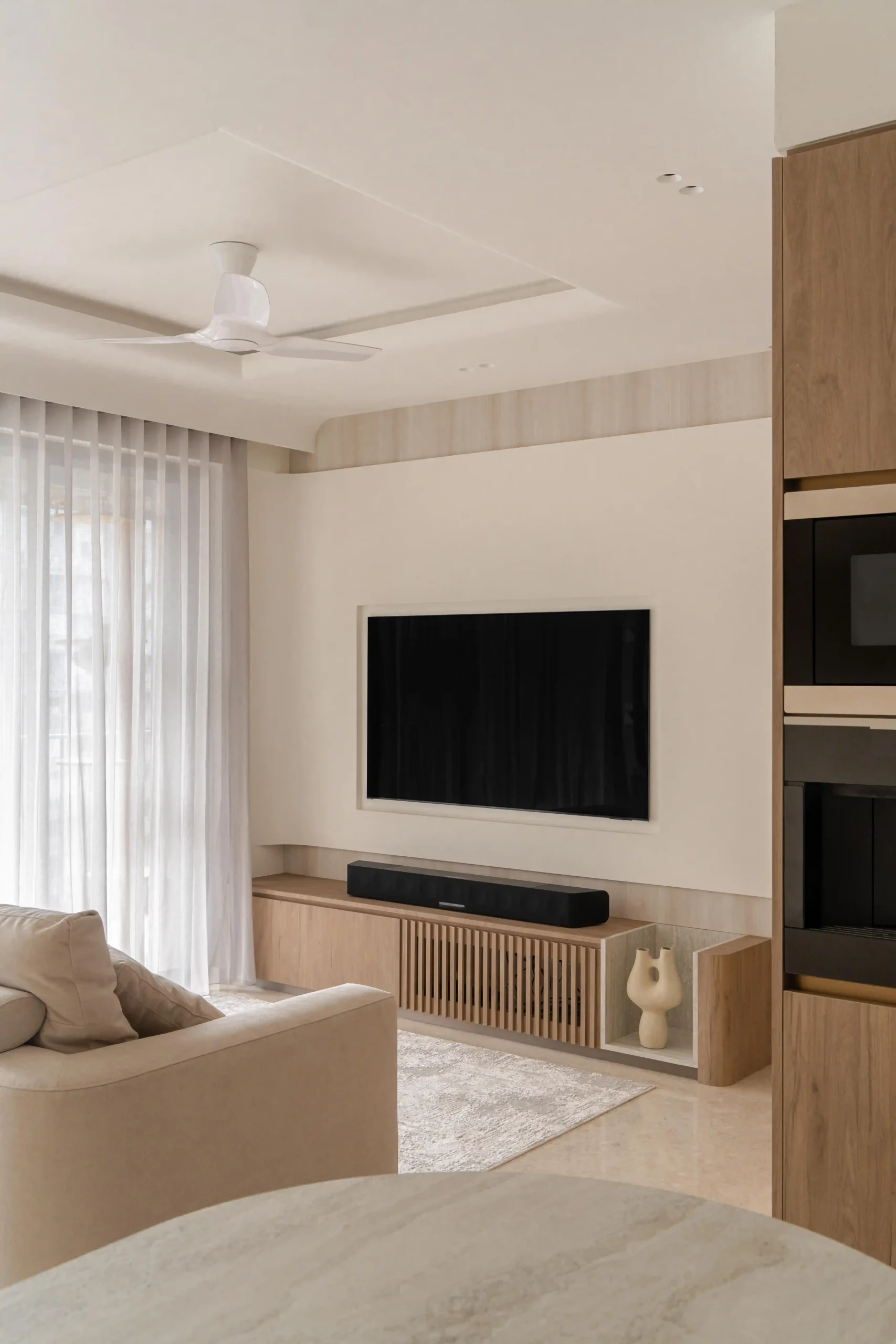 A serene japandi home design living room featuring a wall-mounted TV, slatted wood media console, soft beige sofa, and light-filtering sheer curtains