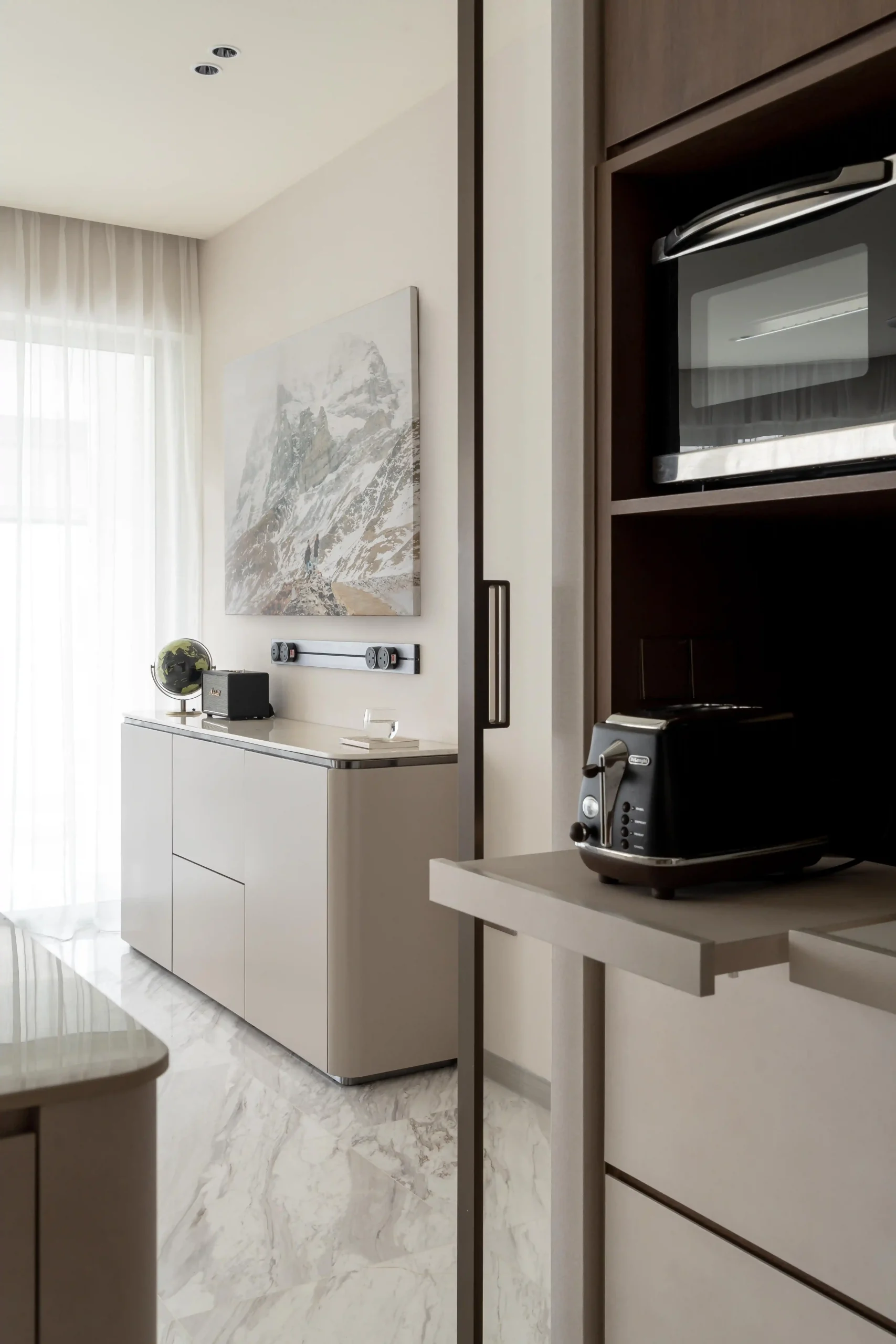 Dry kitchen with matte greige cabinetry and resort-style interior design details — built-in dark wood oven shelving, black De'Longhi toaster, and mountain landscape artwork