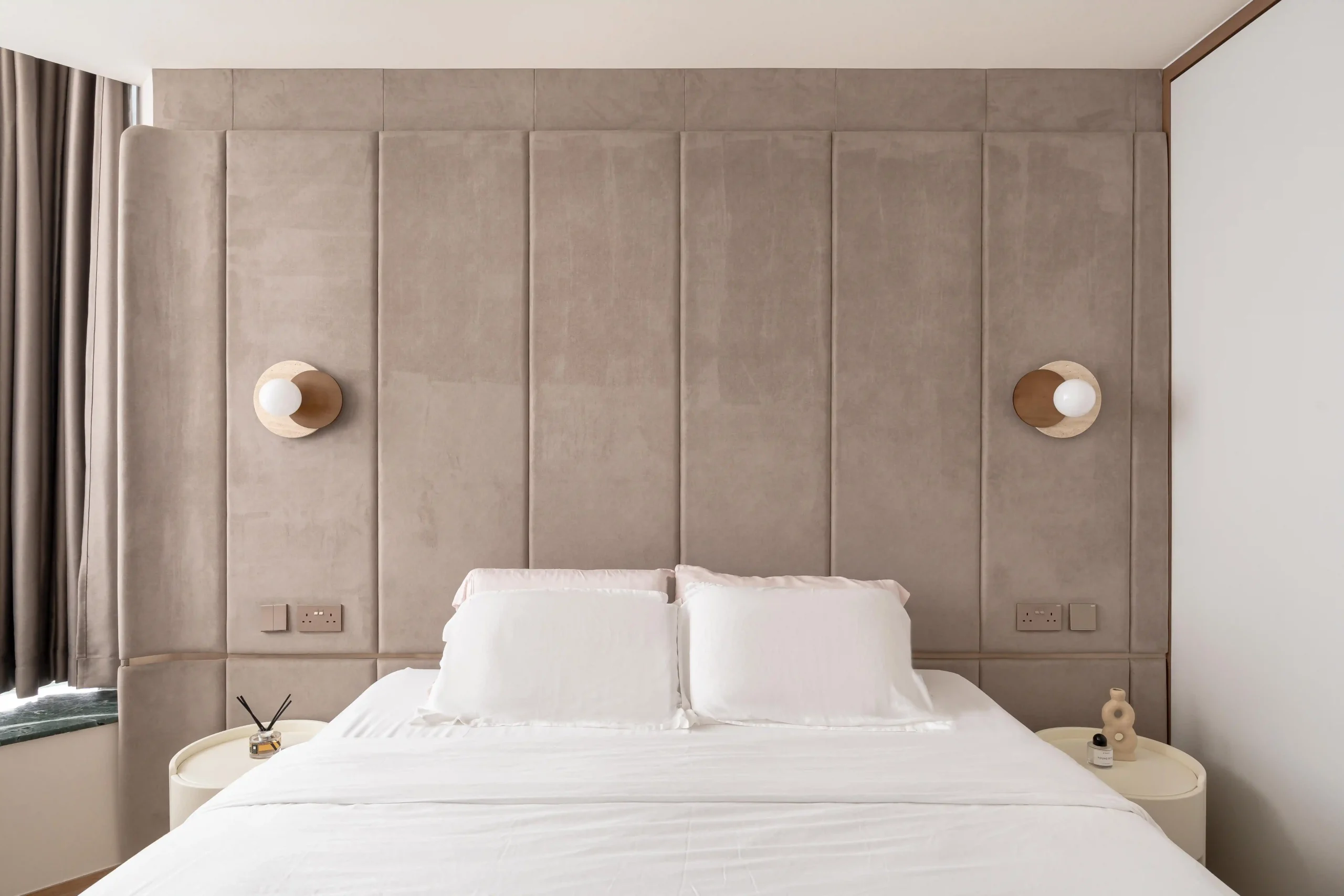 Master bedroom with full-width soft suede upholstered headboard wall, symmetrical travertine globe wall sconces, and crisp white bedding in a resort-style interior design
