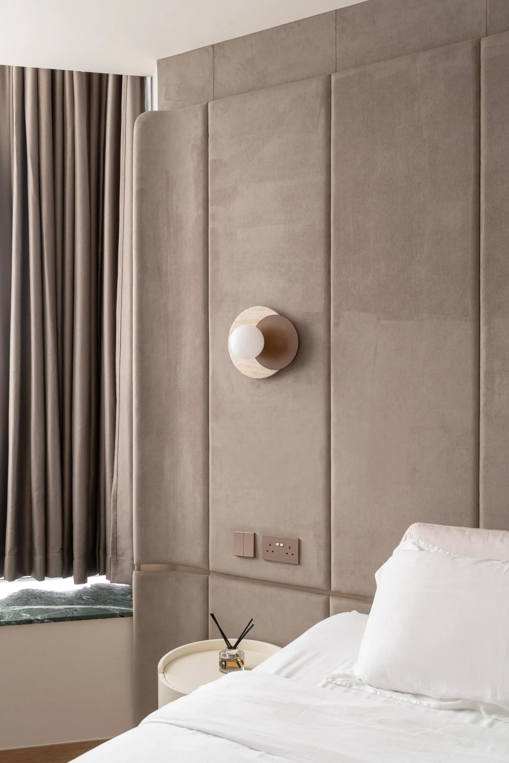 
Floor-to-ceiling suede upholstered headboard wall with travertine globe wall sconce and greige blackout curtains — understated resort-style interior design at its best