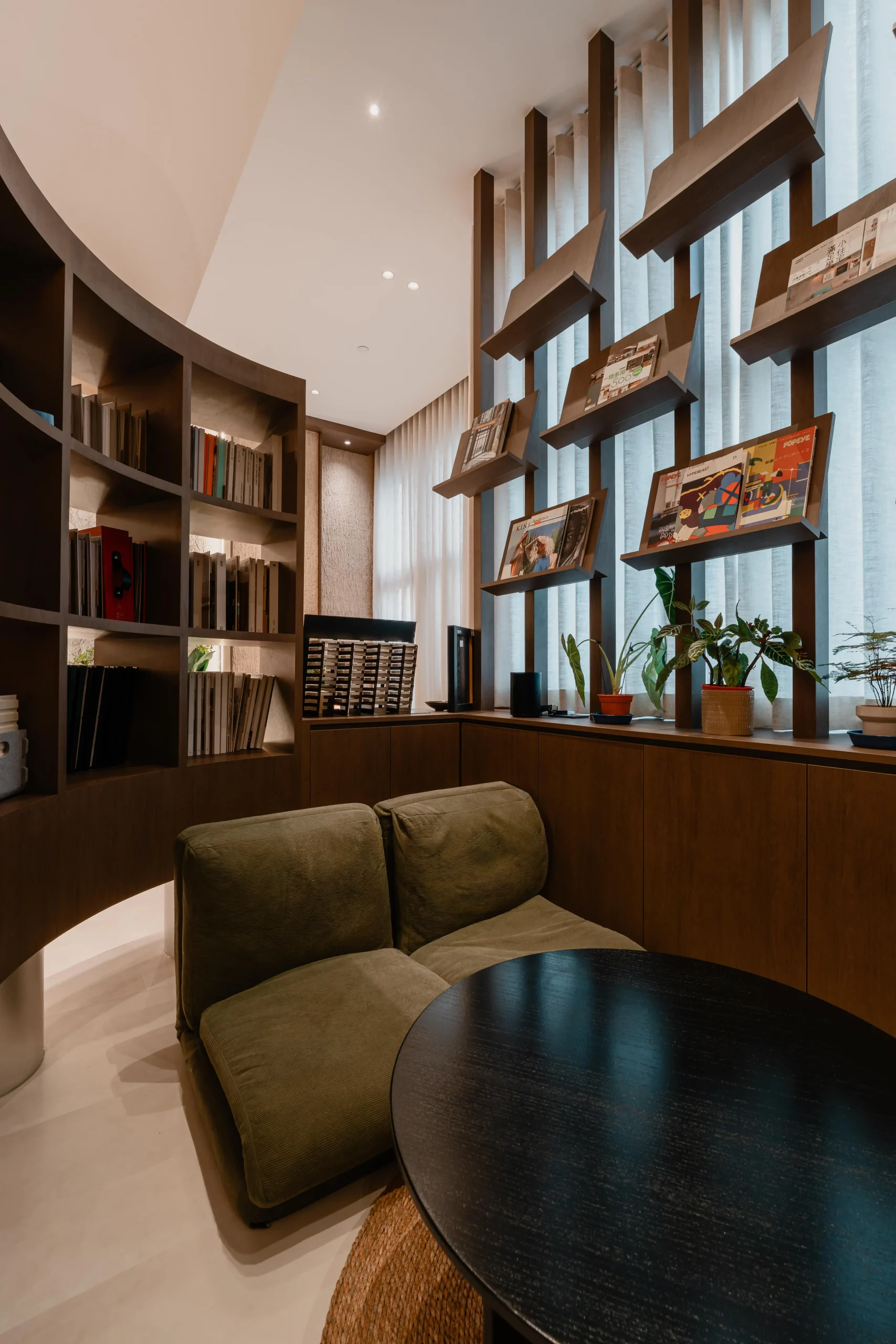 A cozy, private lounge area nestled behind a curved walnut bookshelf with a green modular sofa and black coffee table.
