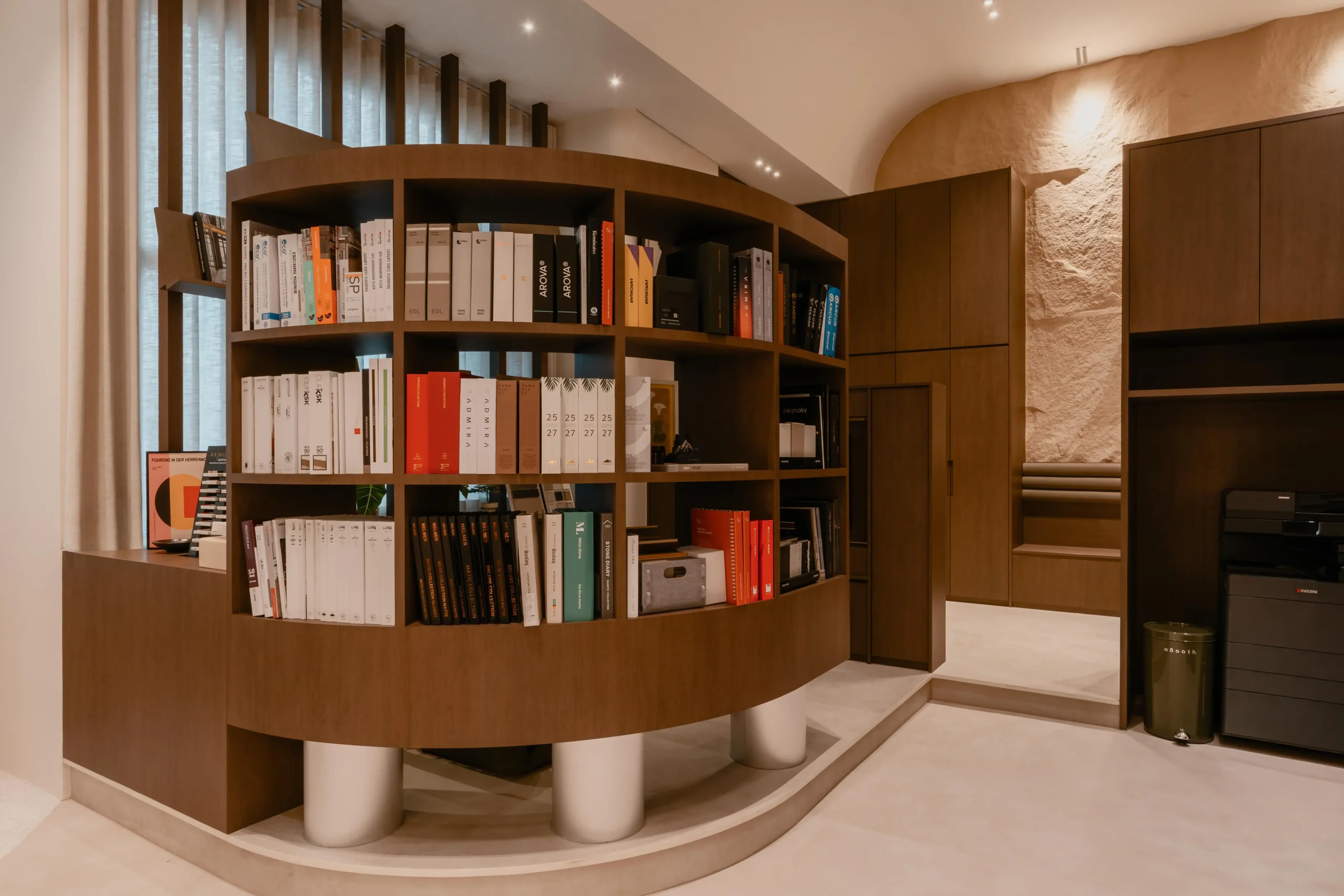 A free-standing architectural curved walnut bookshelf on metal plinth legs inside Sumspace Studio.