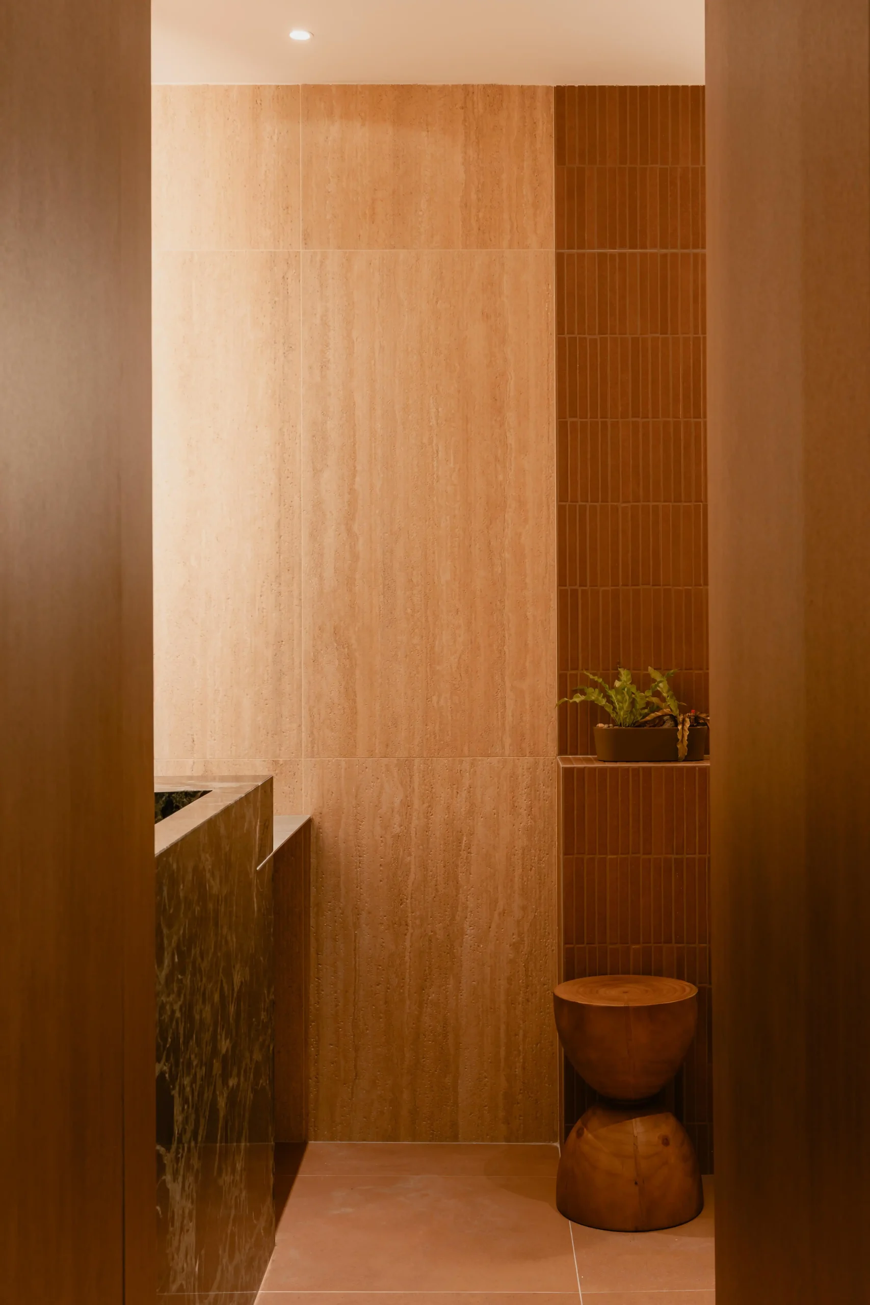 Restroom vanity featuring a dark green marble counter, travertine walls, and a wooden pedestal stool.