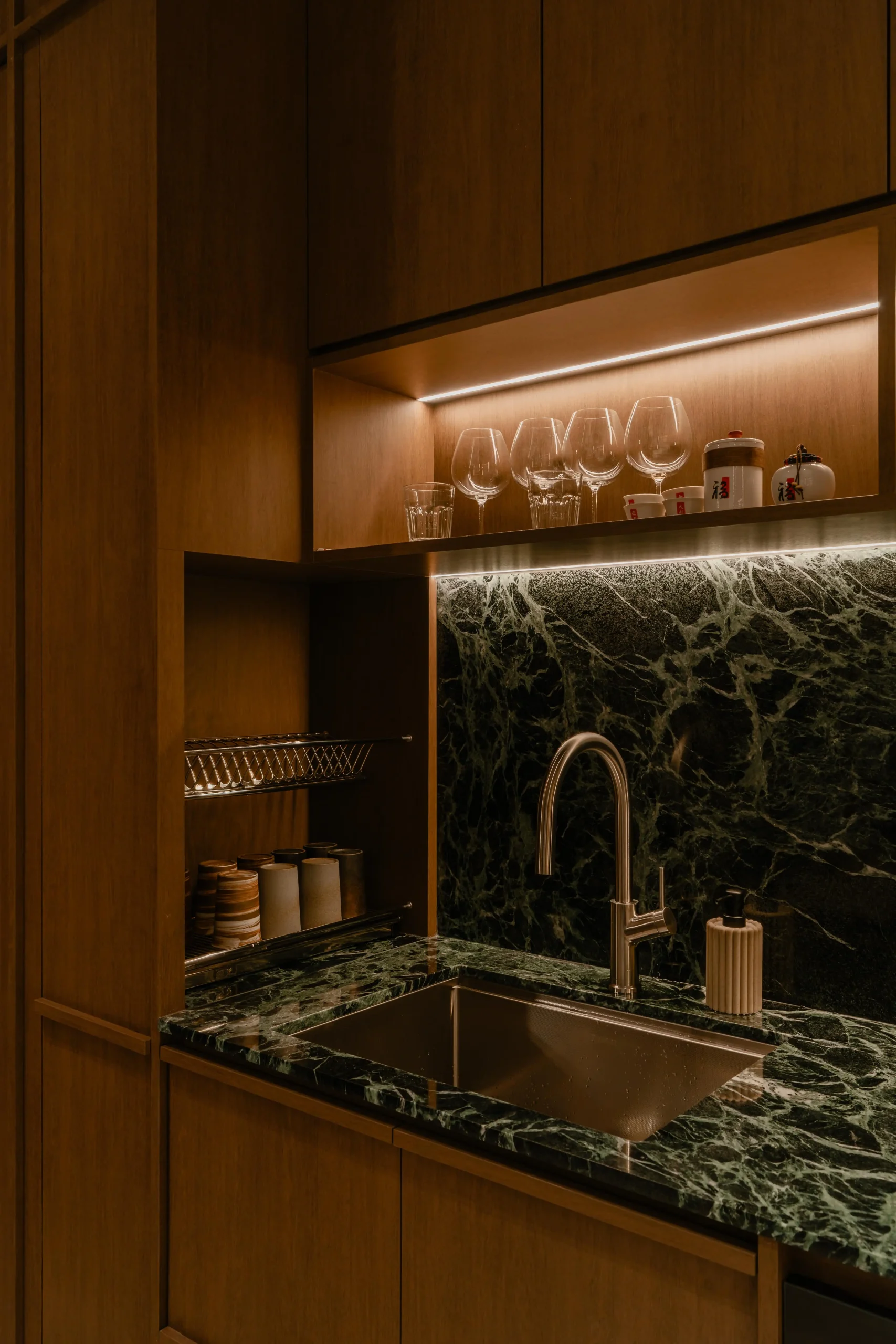 Close-up of the Sumspace Studio pantry showcasing a verde marble backsplash, integrated sink, and illuminated walnut cabinetry.